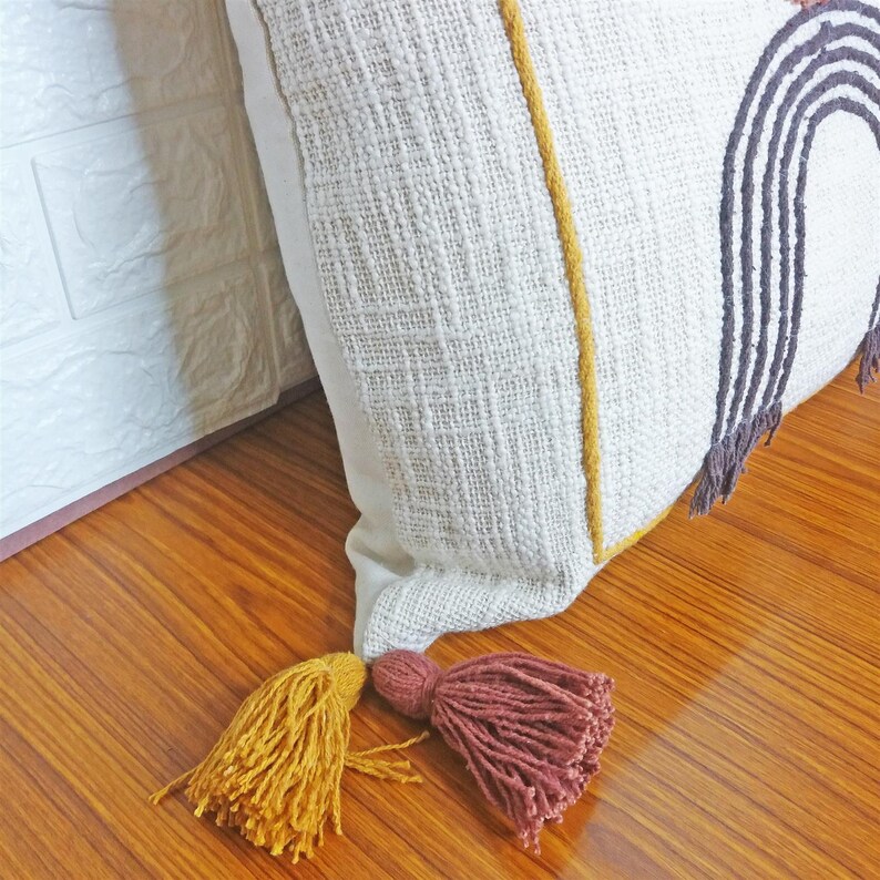 May include: A white throw pillow with a brown and black rainbow design and yellow trim. The pillow has two tassels, one yellow and one brown.