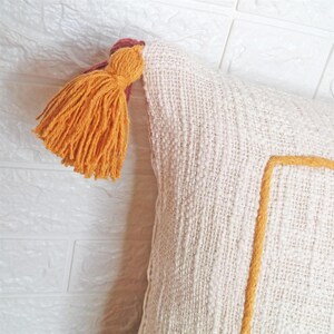 May include: A white throw pillow with a yellow and brown tassel. The pillow has a yellow embroidered square in the center.
