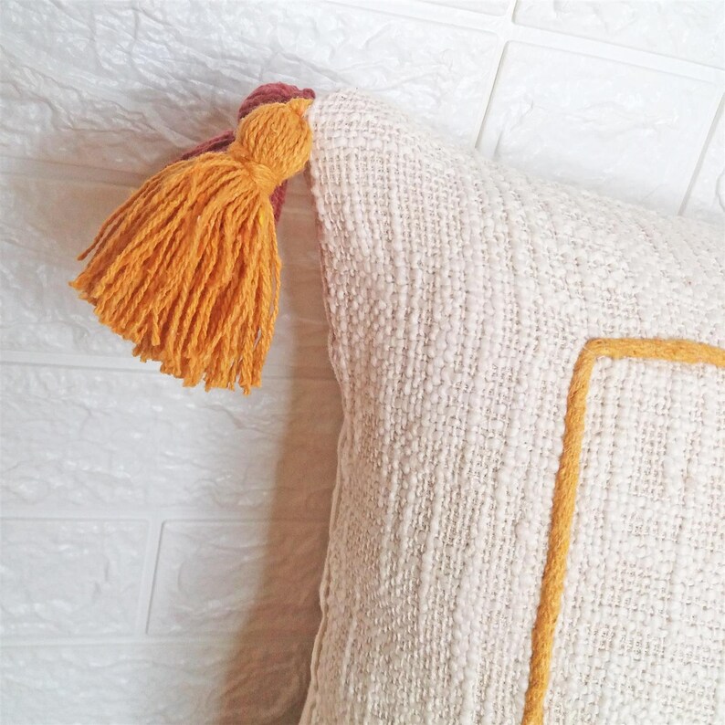 May include: A white throw pillow with a yellow and brown tassel. The pillow has a yellow embroidered square in the center.