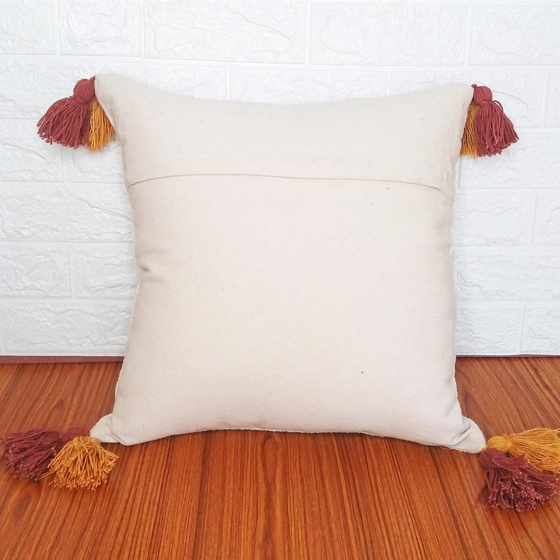 May include: A white throw pillow with tassels in shades of red and yellow. The pillow is made of a soft, natural fabric and has a simple, elegant design.