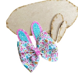 May include: A pink and blue Easter bunny hair bow with a white background and a pattern of pastel colored eggs, flowers, and bunnies.