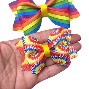 Puede incluir: Dos lazos para el pelo coloridos. Un lazo es de rayas arcoíris y el otro es un patrón tie-dye con remolinos rojos, amarillos, azules y blancos.