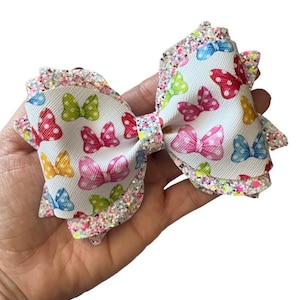 Puede incluir: Un lazo blanco con un borde brillante y un estampado de lazos de colores. El lazo está hecho de tela y tiene un fondo blanco con lazos de colores impresos en él. El lazo está bordeado con un ribete brillante.