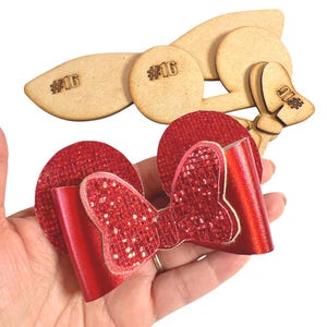 Puede incluir: Orejas de Minnie Mouse rojas con purpurina y un lazo. Las orejas están hechas de tela roja con un acabado brillante. El lazo también es rojo con purpurina y está atado en el centro de las orejas. Las orejas están etiquetadas con el número "#16".