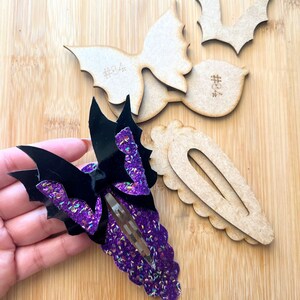 Puede incluir: Pinza para el pelo de murciélago con purpurina morada y negra. La pinza está hecha de purpurina morada y negra y tiene forma de ala de murciélago. La pinza está sobre una superficie de madera.