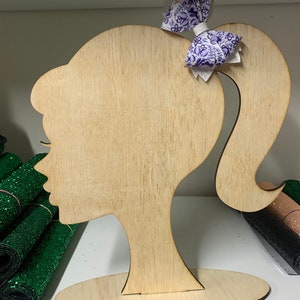Puede incluir: Una silueta de cabeza de muñeca de madera con un lazo floral morado y blanco en la parte superior de la coleta. La cabeza de la muñeca está sobre un soporte de madera.