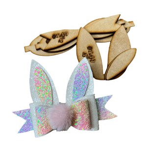 May include: A white and iridescent Easter bunny ear hair bow with a pink pom-pom and wooden cutouts. The bow features glitter accents and a colorful, sparkly design. The wooden cutouts are labeled with the text "#149 4" and are approximately 4 inches long.