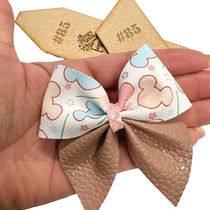 Puede incluir: Un lazo blanco y marrón con estampado de Mickey Mouse. El lazo tiene cabezas de Mickey Mouse rosas, azules y blancas con estrellas y líneas verdes sobre un fondo blanco. El lazo está atado con un material marrón similar al cuero.