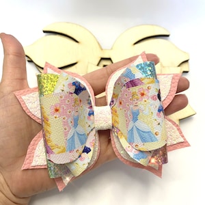May include: A pink, white, and blue layered bow with a princess-themed print. The bow is made of faux leather and has a glittery finish.