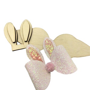 May include: A pink glitter bunny bow with iridescent glitter and a pink pom-pom in the center. The bow is on top of two wooden bunny ear shapes.