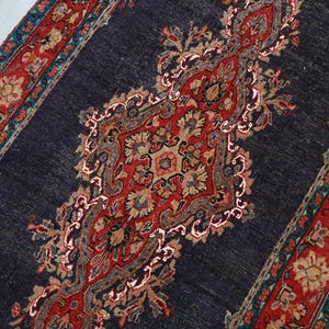 May include: A rectangular, dark blue rug with a central ornate design in red, beige, and gray. The rug features a detailed floral pattern and a red border with teal accents. The rug is likely made of woven material.