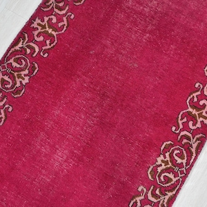 May include: A rectangular, magenta-colored rug with a detailed, repeating floral border in shades of brown and pink. The rug's texture appears slightly worn, adding to its vintage aesthetic. The design is symmetrical.