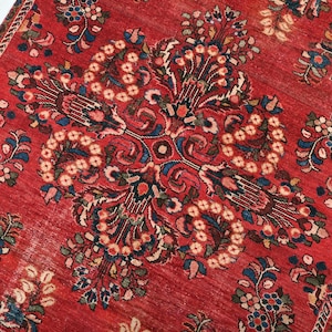 Puede incluir: Primer plano de una alfombra roja con un intrincado diseño floral. La alfombra presenta un medallón central y motivos florales circundantes en tonos azules, verdes y marrones. El borde de la alfombra es de un tono marrón más oscuro.