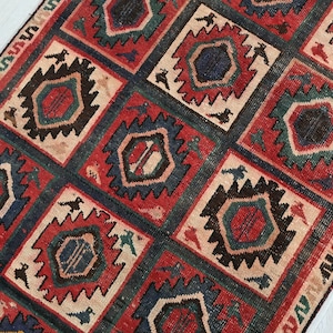May include: A rectangular rug with a repeating geometric pattern. The design features squares with red, blue, and beige elements. Each square contains a central geometric shape and is surrounded by a dark border. The rug's edges have a patterned border.