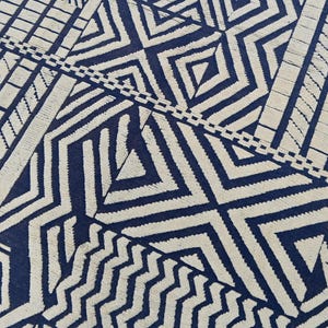 May include: A close-up of a rug with a geometric pattern. The rug features a repeating design of navy blue and off-white shapes, including diamonds, zigzags, and rectangles. The design is symmetrical and covers the entire surface.