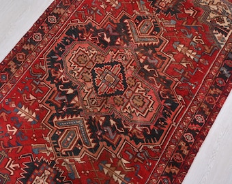 May include: A detailed, rectangular rug with a central geometric design. The rug features a rich red background with intricate patterns in black, brown, and blue. The design includes stylized floral and geometric motifs, creating a traditional, ornate look. The rug's edges are bordered with a repeating pattern.