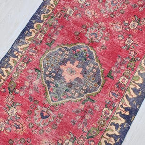 May include: A distressed, rectangular rug with a central medallion design. The rug features a faded red field with a navy blue border and accents of green and beige. The medallion is a mix of blue and peach tones, adding a vintage aesthetic.