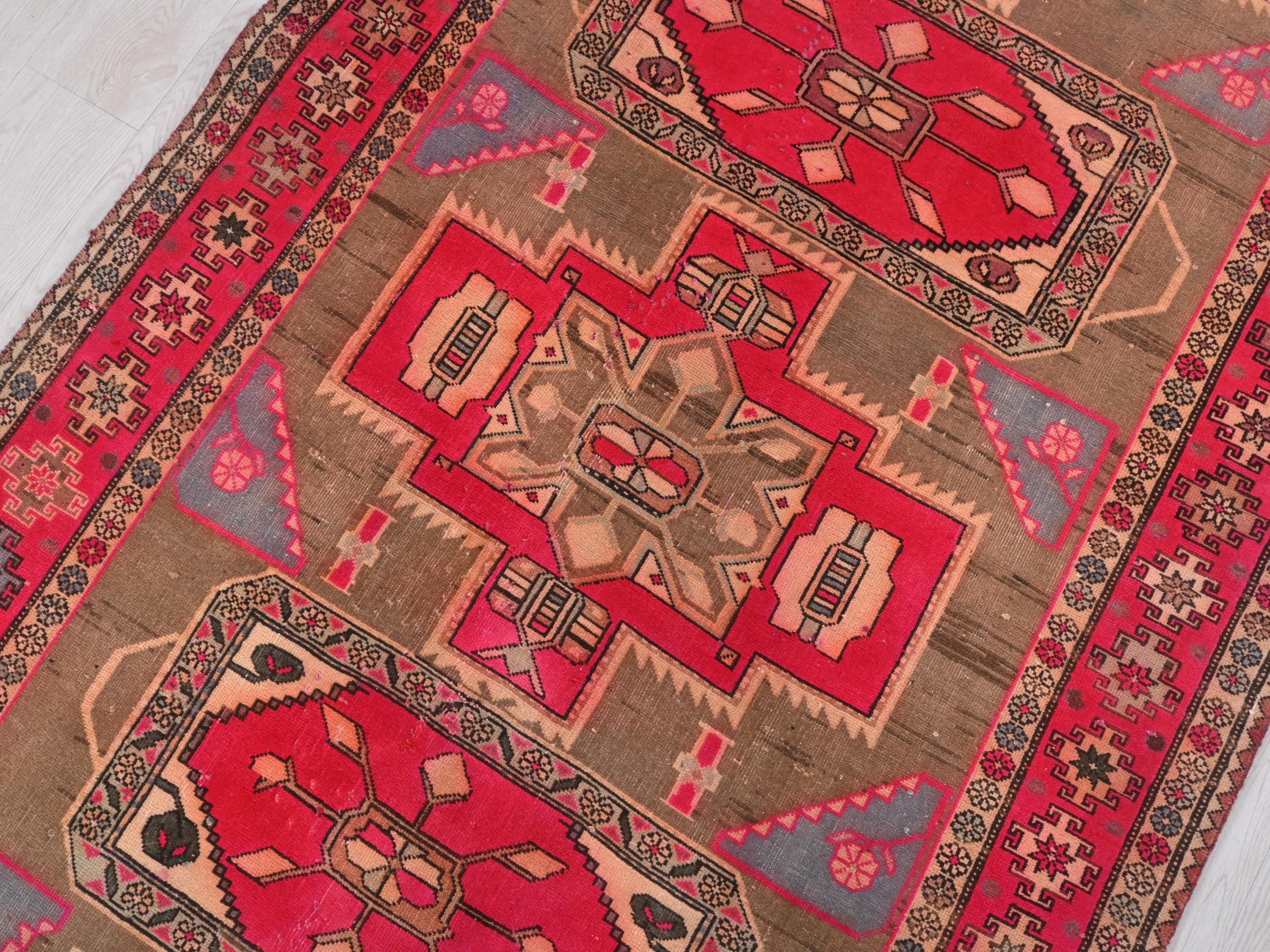 May include: A detailed shot of a vintage rug with a brown and red color scheme. The rug features geometric patterns, including central medallions and border designs. The color palette includes shades of red, brown, beige, and gray.