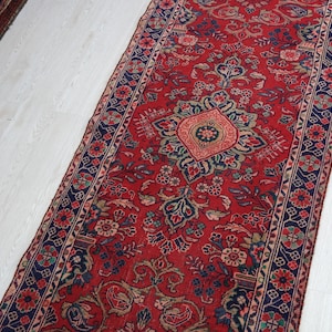 May include: A long rectangular runner rug featuring a vibrant red background with intricate floral and geometric patterns in navy blue, light blue, and cream. The design includes a central medallion motif surrounded by dense botanical elements.