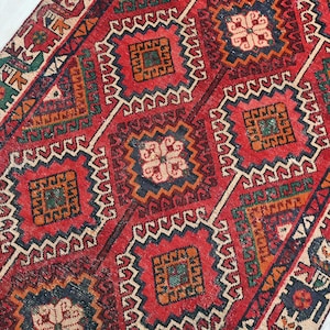 Puede incluir: Una alfombra vintage detallada con un patrón geométrico repetitivo. La alfombra presenta un fondo rojo con diseños en forma de diamante en tonos de azul, verde, naranja y crema. Los intrincados patrones y los colores ricos crean una estética clásica y atemporal.