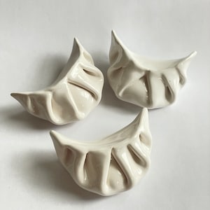 Peut inclure: Trois raviolis en c&eacute;ramique blanche, chacun avec une forme et une texture uniques, sont dispos&eacute;s sur un fond blanc. Les raviolis sont un objet d&eacute;coratif et ne sont pas destin&eacute;s &agrave; &ecirc;tre consomm&eacute;s.