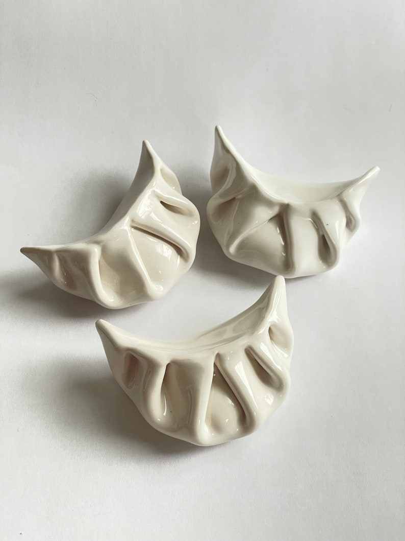 Peut inclure: Trois raviolis en c&eacute;ramique blanche, chacun avec une forme et une texture uniques, sont dispos&eacute;s sur un fond blanc. Les raviolis sont un objet d&eacute;coratif et ne sont pas destin&eacute;s &agrave; &ecirc;tre consomm&eacute;s.