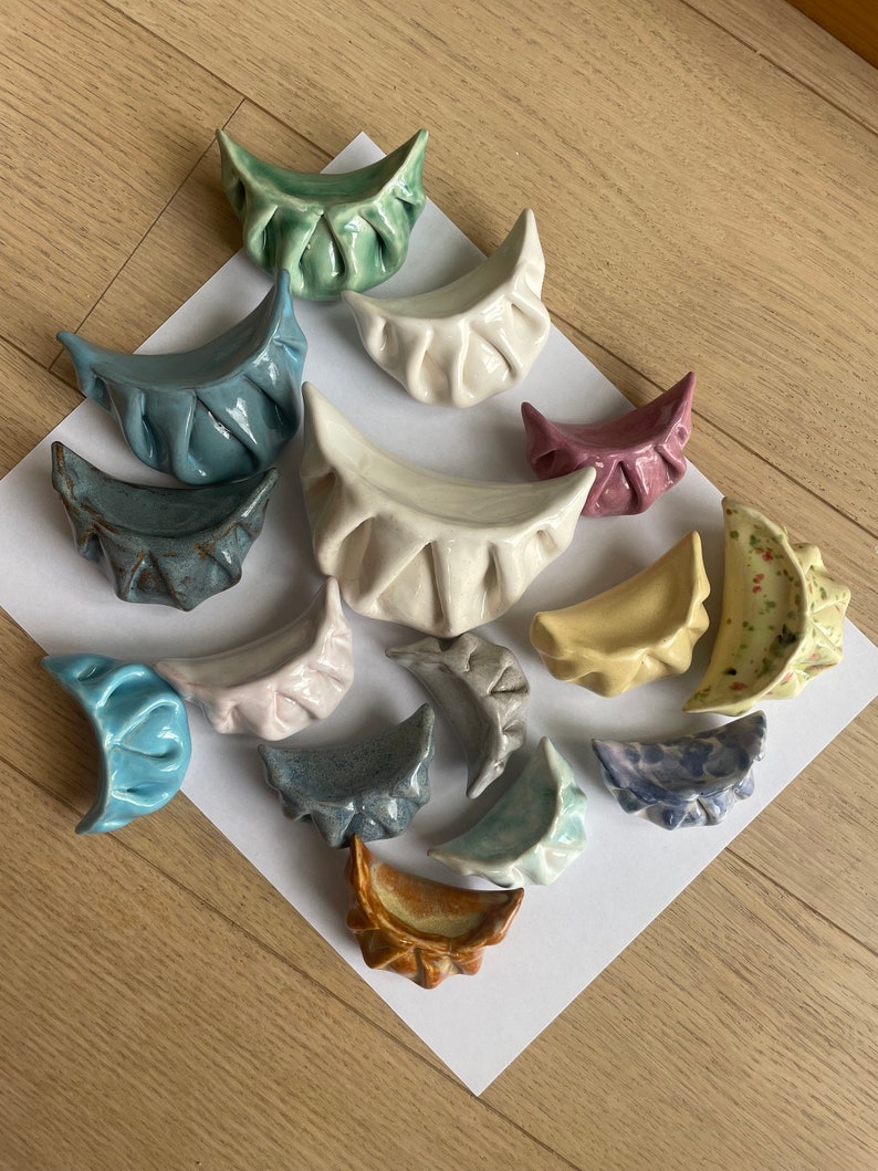 Peut inclure: Une collection de 15 raviolis en c&eacute;ramique de diff&eacute;rentes couleurs et motifs, notamment bleu, vert, jaune, marron et blanc. Les raviolis sont dispos&eacute;s sur une surface blanche.