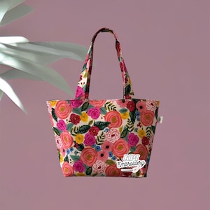May include: A floral tote bag with a cream background and a vibrant pattern of pink, yellow, and red roses, green leaves, and blue accents. The bag has matching handles and the text "KPI Embroidery".