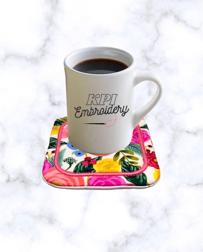 May include: A white ceramic mug filled with black coffee, featuring the black text KPI Embroidery and a small needle graphic, resting on a square fabric coaster with a vibrant floral pattern of pink, yellow, and green on a white background.