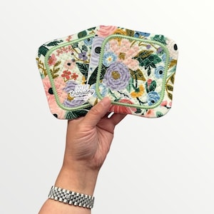 May include: Two square quilted fabric coasters featuring a floral pattern with pink, blue, yellow, and green flowers on a white background. The edges are finished with light green piping. The text KPI Embroidery is printed on the front surface.