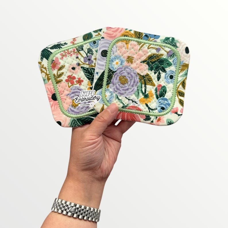 May include: Two square quilted fabric coasters featuring a floral pattern with pink, blue, yellow, and green flowers on a white background. The edges are finished with light green piping. The text KPI Embroidery is printed on the front surface.