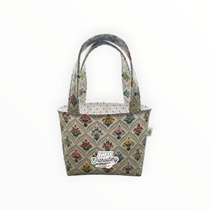 May include: A small fabric tote bag featuring a cream-colored exterior with a repeating diamond pattern containing colorful floral motifs. The interior lining is white with black polka dots. The text KPI Embroidery is printed on the front.