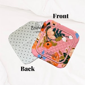 May include: A square quilted pot holder featuring a pink background with a floral pattern of orange, white, and blue flowers. The reverse side is light gray with small black polka dots. The text KPI Embroidery is visible on both sides.