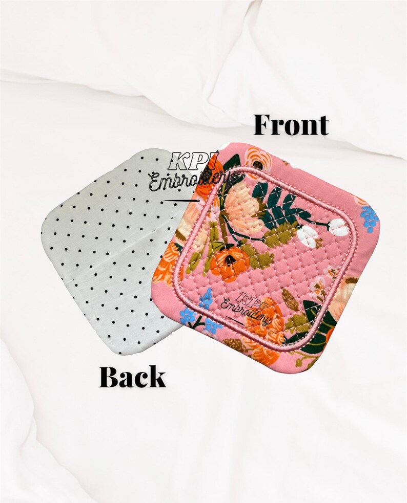 May include: A square quilted pot holder featuring a pink background with a floral pattern of orange, white, and blue flowers. The reverse side is light gray with small black polka dots. The text KPI Embroidery is visible on both sides.