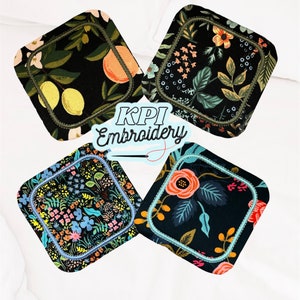 May include: Four square fabric coasters with rounded corners, each featuring a distinct black-based floral or fruit print. The coasters are outlined with decorative machine embroidery stitching. The text KPI Embroidery is centered in the image.
