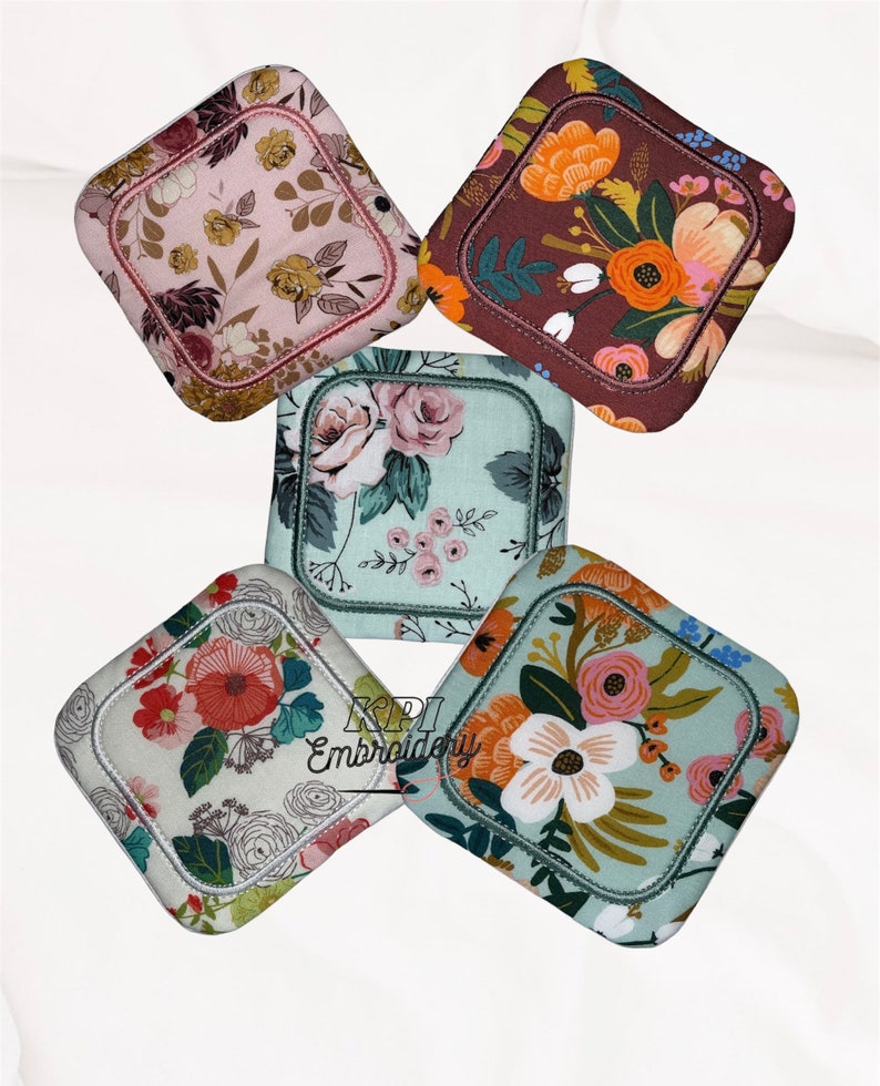 May include: Five square fabric coasters with rounded corners, each featuring distinct floral patterns in shades of pink, brown, orange, and light blue. The text KPI Embroidery is printed in black over the bottom left coaster.
