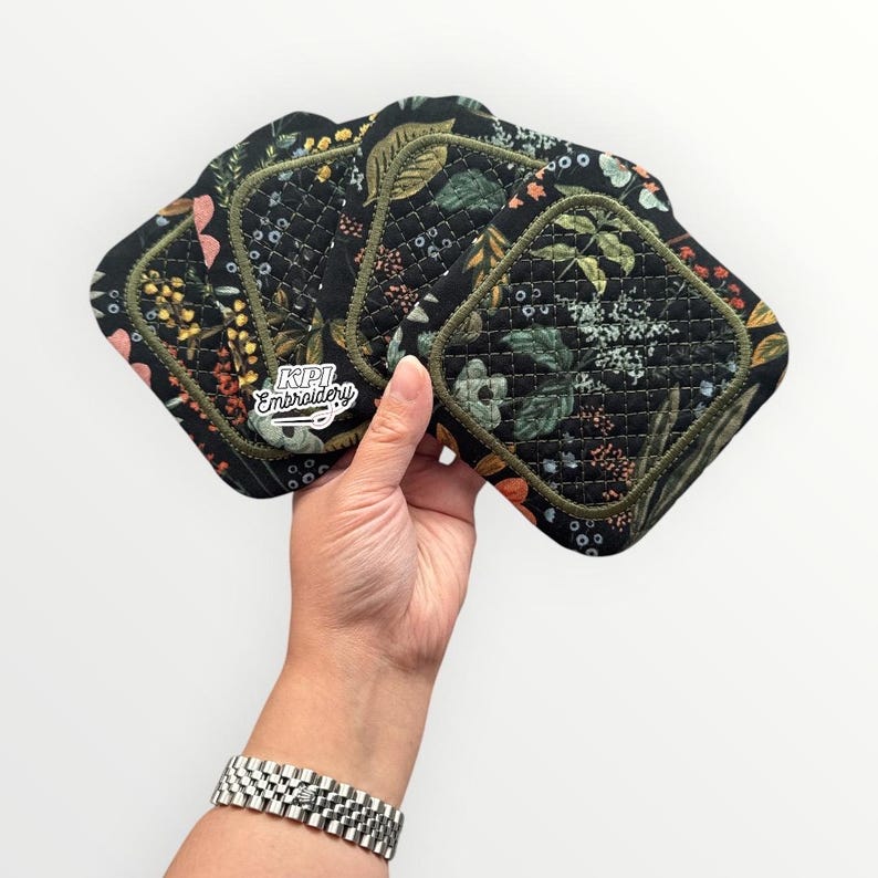 May include: A set of four square quilted fabric coasters featuring a black background with a colorful botanical print of leaves, flowers, and berries. Each coaster has a dark olive green stitched border and the text KPI Embroidery is visible.