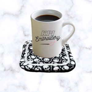 May include: A white ceramic mug filled with dark liquid, sitting on a black and white skull-patterned coaster. The mug features the text "KPI Embroidery". The coaster has a white border.