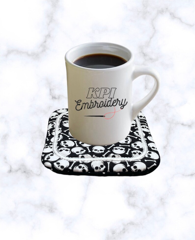 May include: A white ceramic mug filled with dark liquid, sitting on a black and white skull-patterned coaster. The mug features the text "KPI Embroidery". The coaster has a white border.