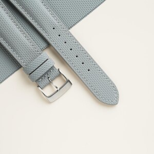 Puede incluir: Una correa de reloj gris claro con una superficie texturizada y costuras blancas. La correa presenta una hebilla de color plateado y se muestra sobre un fondo blanco. La correa del reloj est&aacute; hecha de un material duradero.