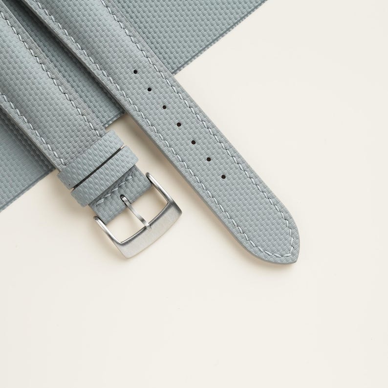 Puede incluir: Una correa de reloj gris claro con una superficie texturizada y costuras blancas. La correa presenta una hebilla de color plateado y se muestra sobre un fondo blanco. La correa del reloj est&aacute; hecha de un material duradero.