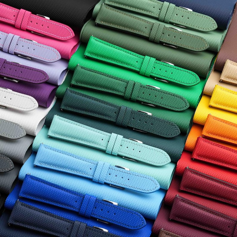 May include: A collection of colorful watch straps in various shades, including pink, purple, green, blue, and yellow. The straps are made of a textured material and feature silver-tone buckles. The arrangement showcases a spectrum of colors.