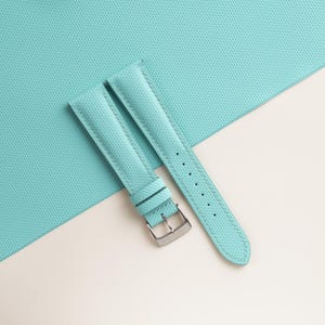 Puede incluir: Una correa de reloj azul claro con una superficie texturizada y una hebilla plateada. La correa est&aacute; hecha de un material duradero y presenta costuras en los bordes. El fondo es una superficie texturizada azul claro.