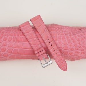 May include: A pair of pink watch straps with a crocodile skin texture, featuring a silver-colored buckle. The straps are laid over a matching pink crocodile skin surface. The straps are designed for a watch.