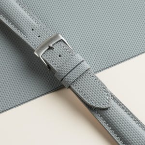 Puede incluir: Primer plano de una correa de reloj gris con una superficie texturizada y costuras blancas. La correa presenta una hebilla plateada y se encuentra sobre un fondo gris claro y blanco. Un elegante accesorio para relojes.