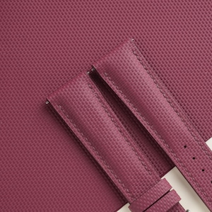 May include: Close-up of a burgundy watch strap with a textured surface. The strap is made of a durable material and features detailed stitching. The background is a matching burgundy material.