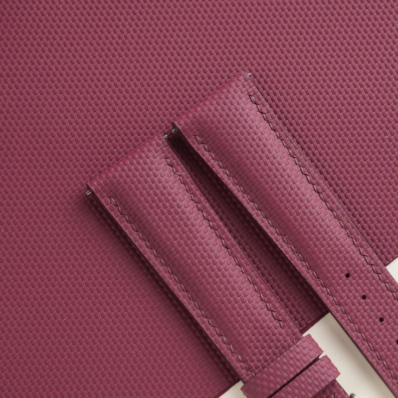 May include: Close-up of a burgundy watch strap with a textured surface. The strap is made of a durable material and features detailed stitching. The background is a matching burgundy material.