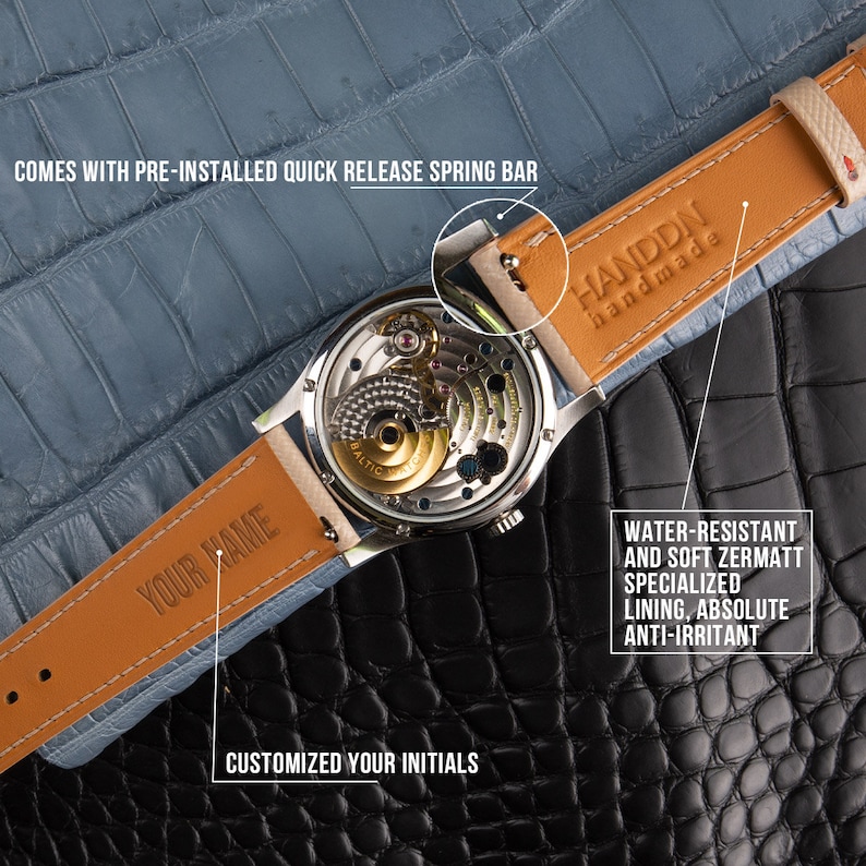 May include: A close-up of a silver wristwatch with a transparent back, showcasing the intricate internal mechanisms. The watch has a tan leather strap with blue lining and the text "YOUR NAME". The strap is labeled "HANDDN handmade". The text "COMES WITH PRE-INSTALLED QUICK RELEASE SPRING BAR" is also visible.