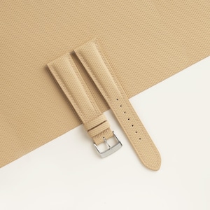 Puede incluir: Correa de reloj de color beige con superficie texturizada y hebilla plateada. La correa marr&oacute;n claro tiene costuras en los bordes. La correa est&aacute; colocada en diagonal sobre un fondo beige y blanco.