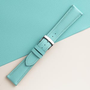 Puede incluir: Una correa de reloj turquesa con superficie texturizada y costuras blancas. Cuenta con una hebilla plateada y se muestra sobre un fondo turquesa claro texturizado.